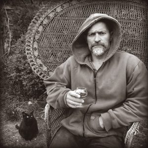 Black and white photo of Omar Rayyan wearing a hooded jacket, holding a wine glass, seated in a peacock-style wicker chair, black cat in background
